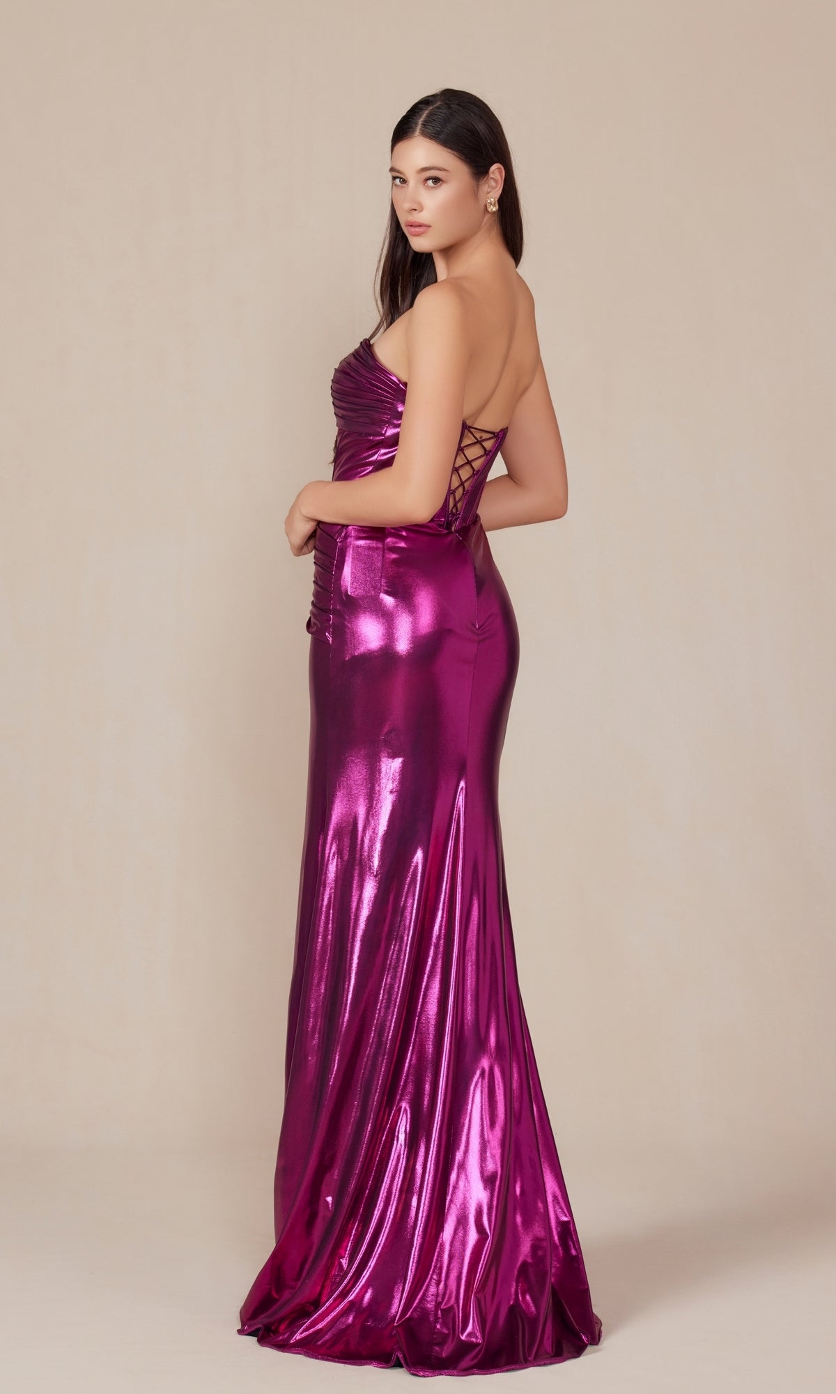 A woman models the Pleated Metallic Long Prom Dress: Nox Anabel T1499, featuring a shiny magenta hue and lace-up corset back, set against a plain beige background.